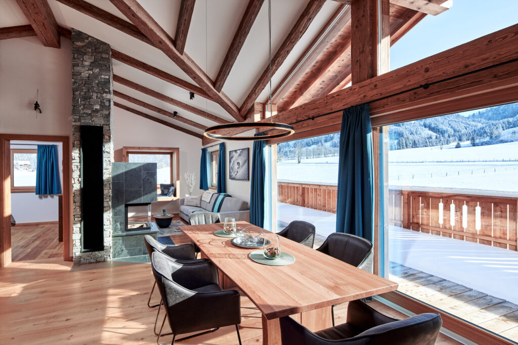 Geräumiges Wohnzimmer in einem Chalet mit Holzbalken, einem großen Esstisch, blauen Vorhängen und Blick auf winterliche Landschaft.